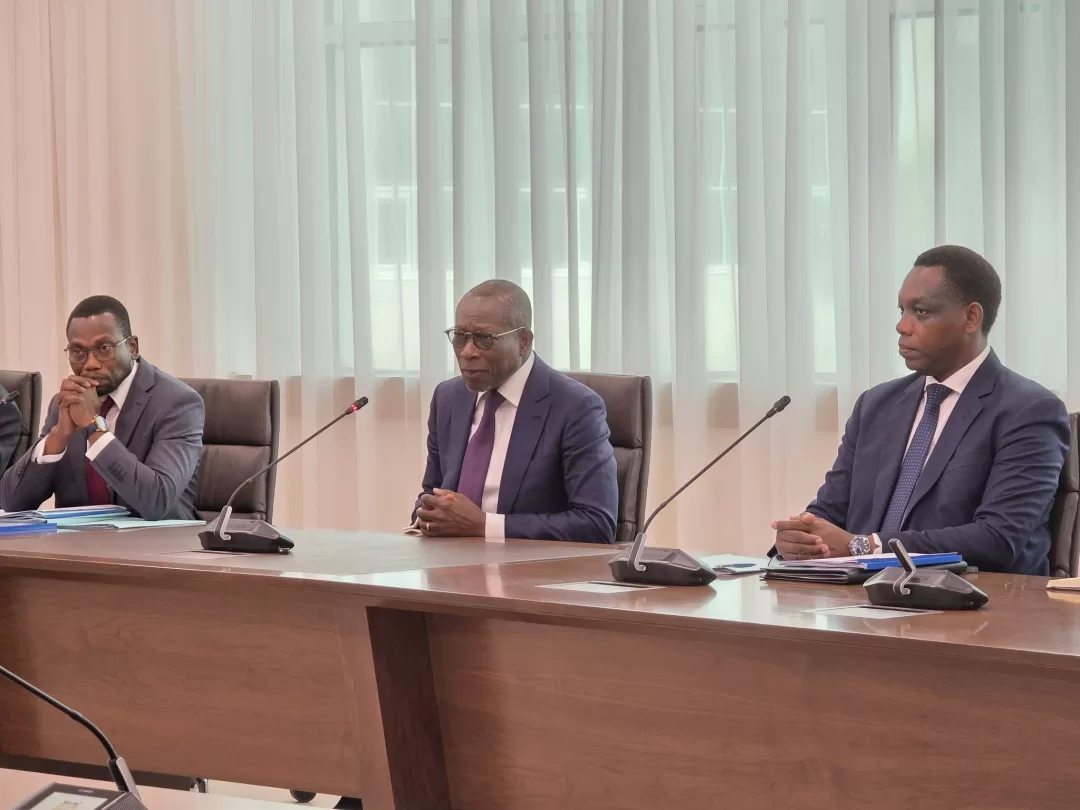 Trois hommes en costume siègent à une table de réunion avec des microphones devant eux dans une salle lumineuse