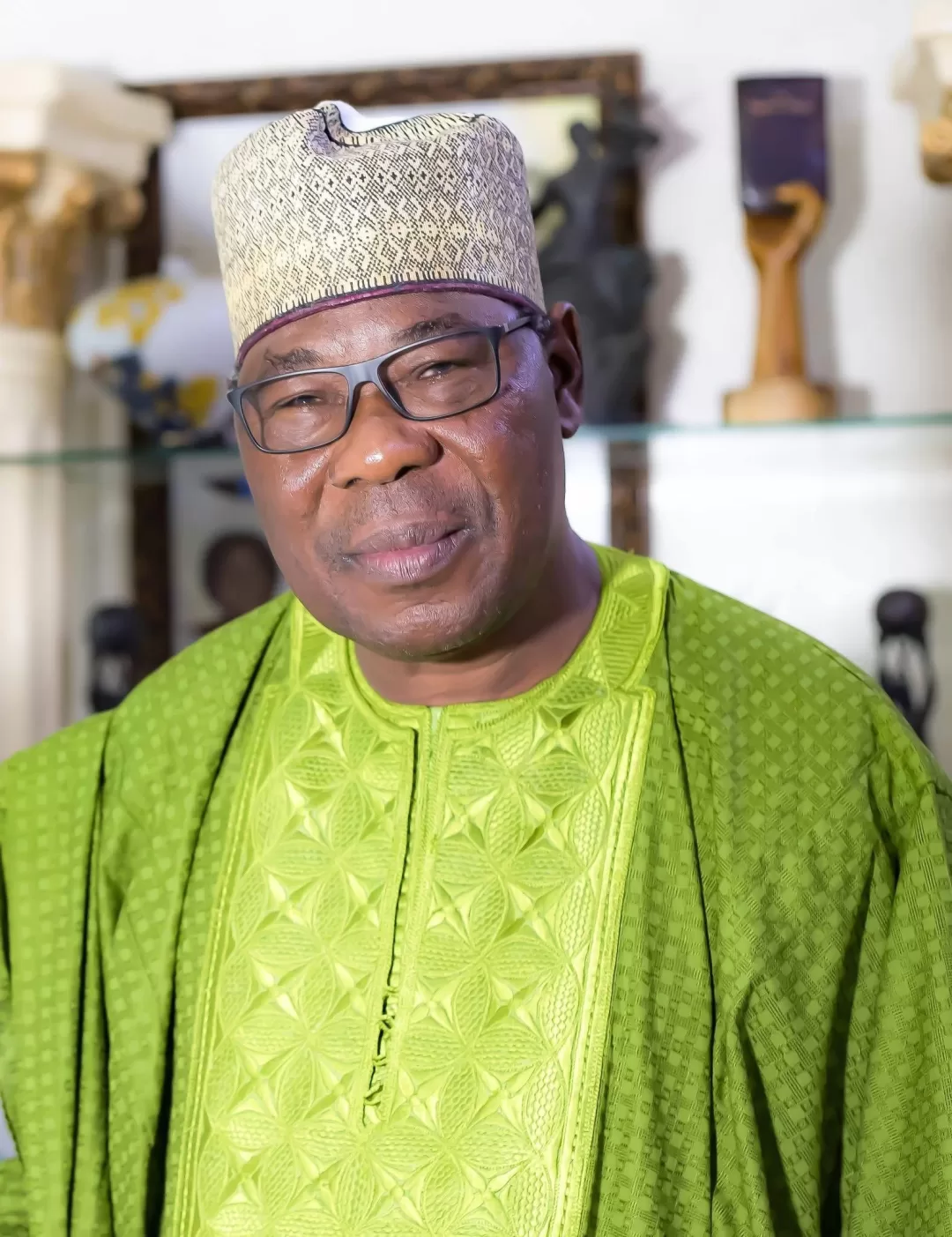 Homme portant un boubou vert et un chapeau traditionnel beige avec des lunettes debout devant une étagère avec objets décoratifs
