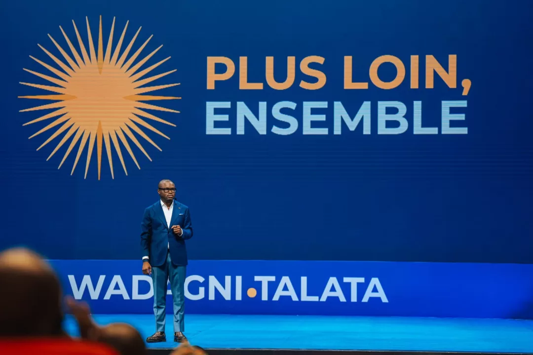 Un homme en costume debout sur scène devant un grand écran bleu avec le texte « Plus loin, ensemble