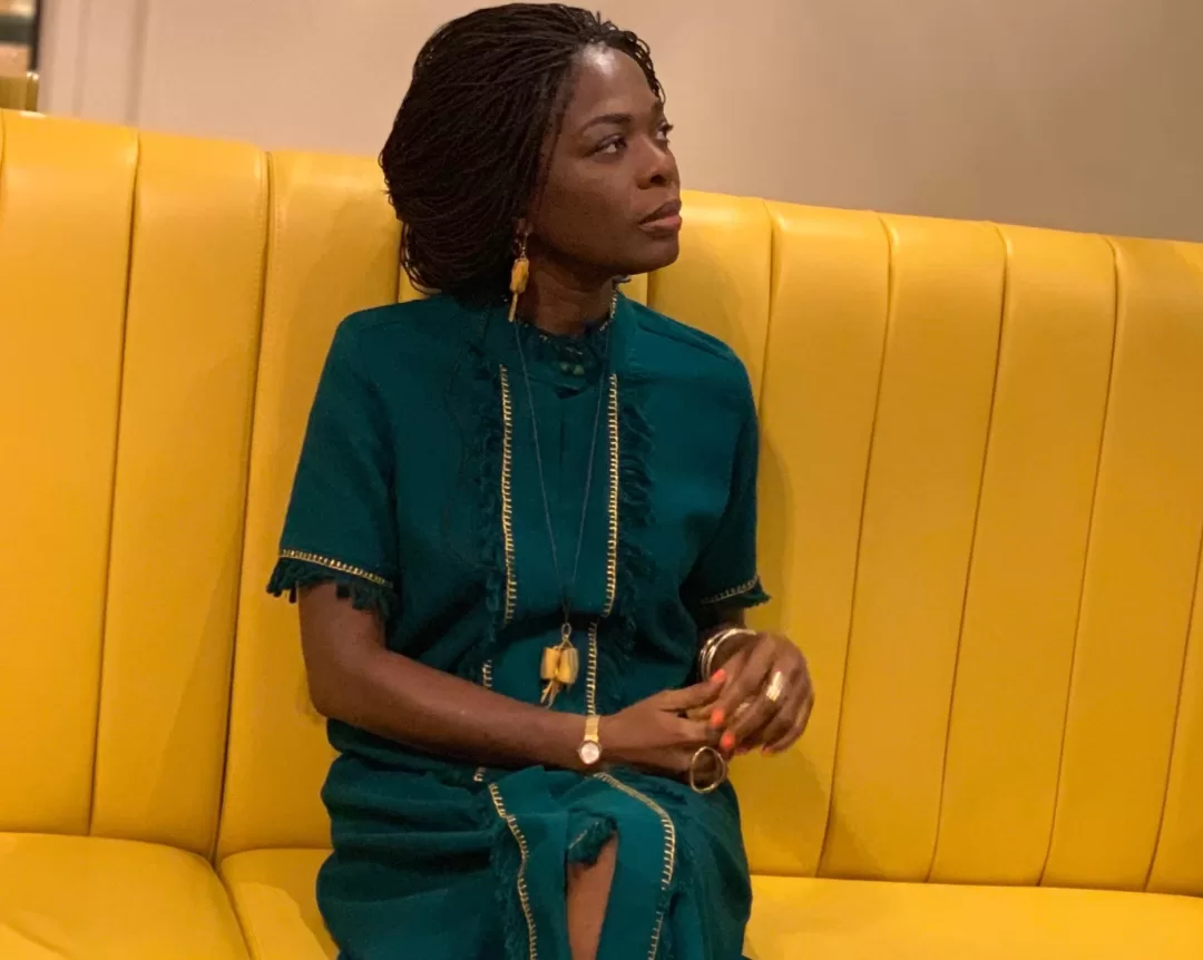 Femme assise sur un canapé jaune regardant vers la droite vêtue d’une robe verte avec des bijoux dorés