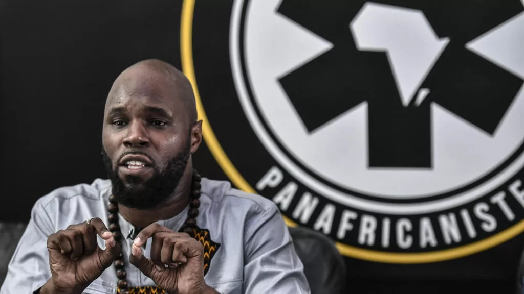 Un homme barbu en chemise grise gesticule devant un logo rond noir et blanc avec un continent africain stylisé