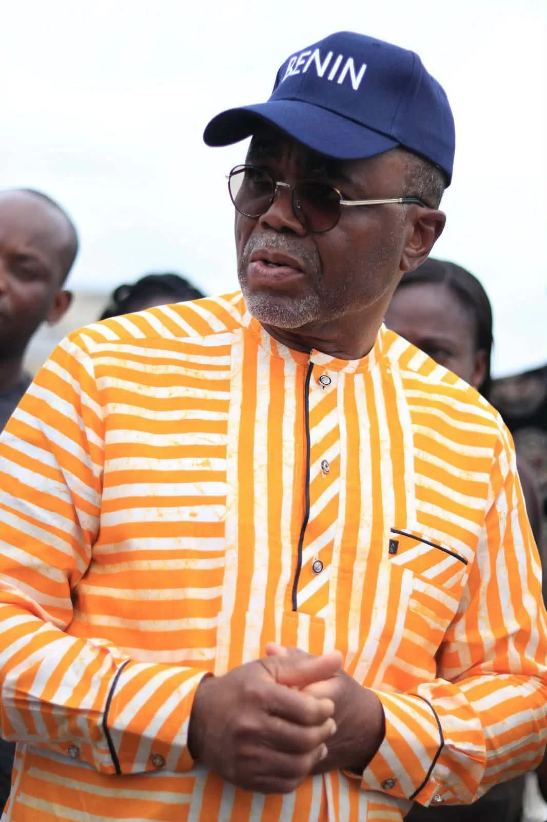 Un homme portant une chemise rayée orange et blanche, une casquette bleue et des lunettes regarde vers la droite avec inquiétude