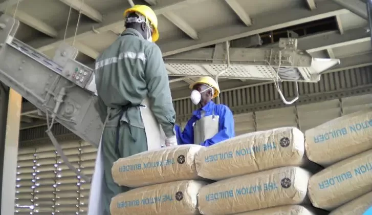 Deux ouvriers en tenue de protection et casque discutent près de sacs de ciment empilés dans une usine