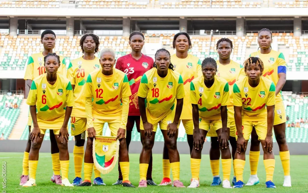 Onze joueuses de football en tenue jaune posent sur un terrain dans un stade vide assis sur des gradins multicolores