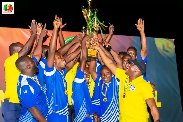 Des joueurs en maillots bleus et un encadrant en jaune lèvent ensemble un trophée de champion sous les acclamations