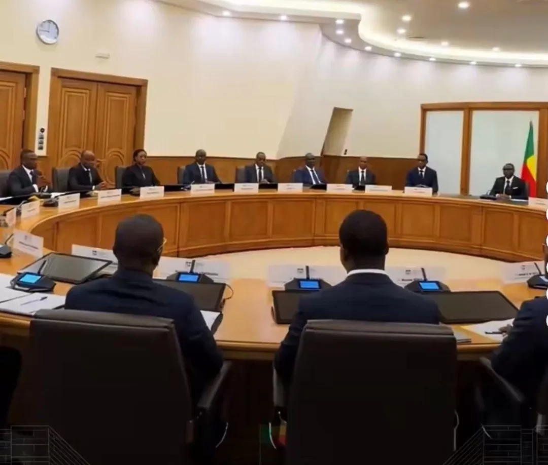 Des personnes en costume assises autour d'une grande table ovale dans une salle de réunion officielle avec un drapeau dans le coin