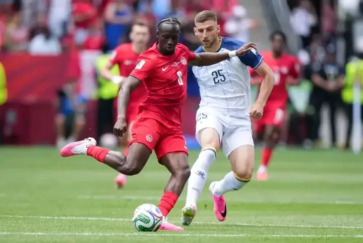 Deux joueurs de football en pleine action, l'un en tenue rouge frappant le ballon, l'autre en blanc le défiant sur le terrain vert