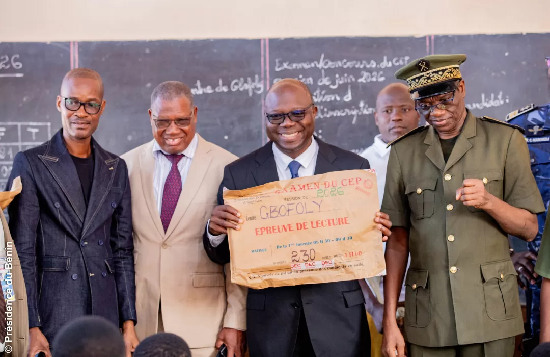 Quatre hommes souriants dont un en uniforme posent devant un tableau noir avec un grand papier indiquant une épreuve de lecture
