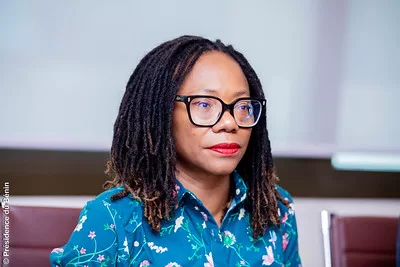 Une femme aux cheveux tressés et lunettes noires porte une chemise bleue à motifs floraux assise devant un mur clair
