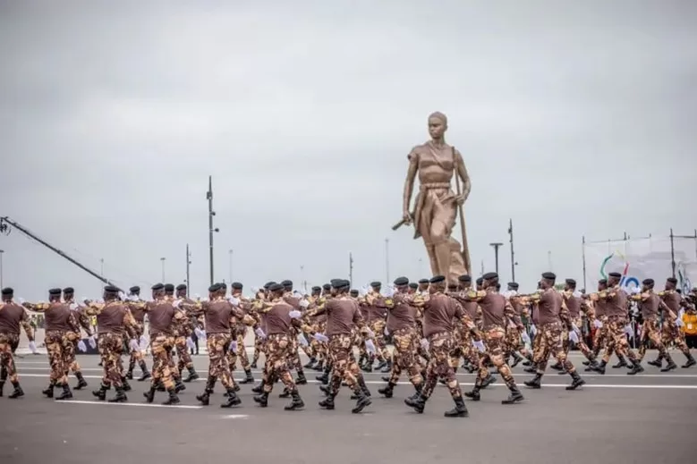 Des militaires en uniforme marron et camouflage marchent en formation devant une grande statue humaine en bronze sur une place ouverte