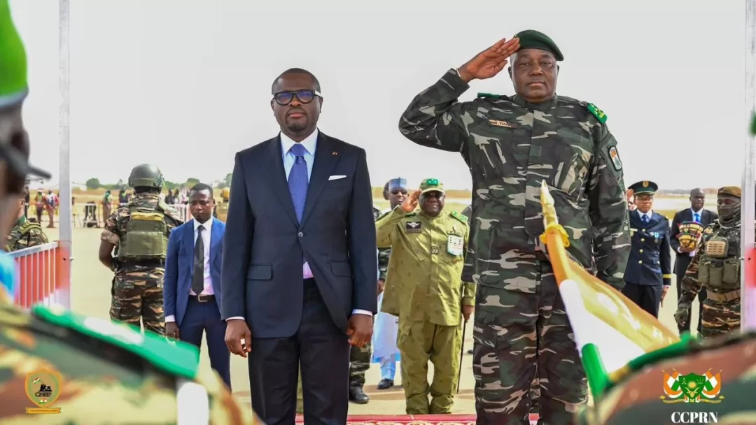 Un homme en costume sombre et un militaire saluent debout devant des soldats en uniformes face à eux
