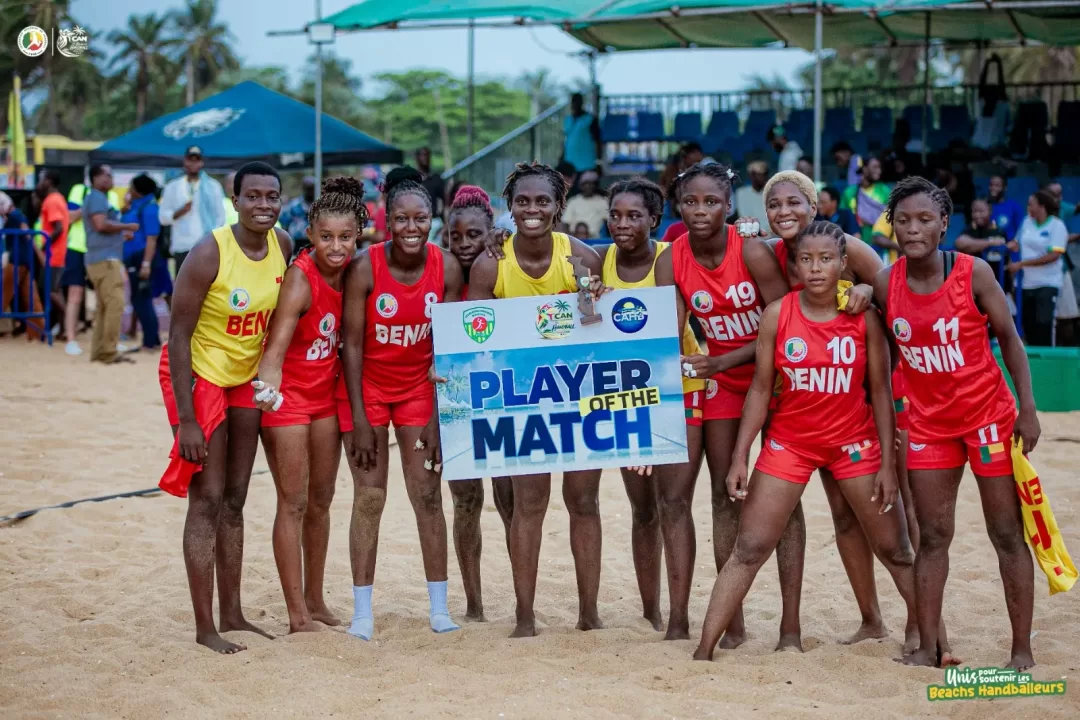 Dix joueuses de beach handball posent ensemble sur le sable avec un panneau « Player of the Match » devant des spectateurs assis et debout