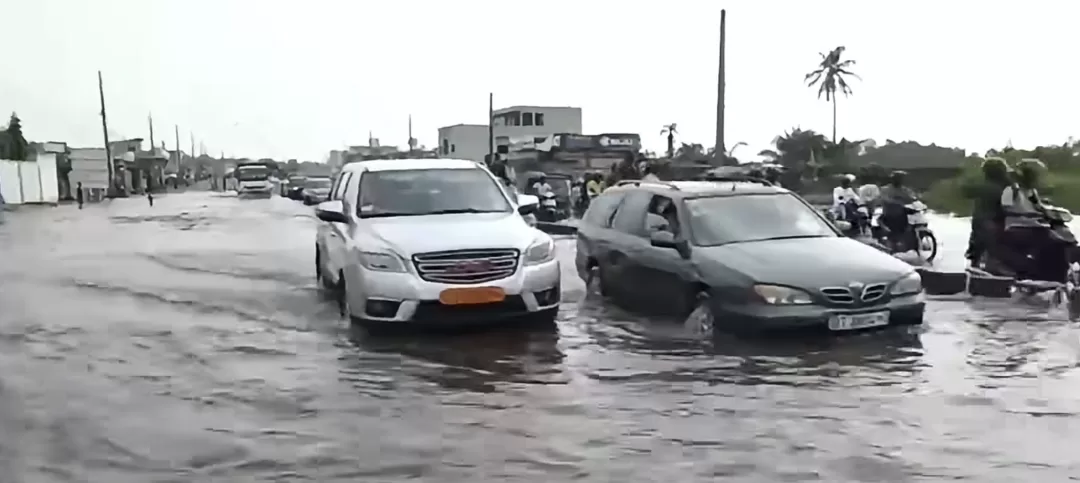 Voitures circulant dans une rue inondée avec des motos et des piétons sur le côté sous un ciel gris