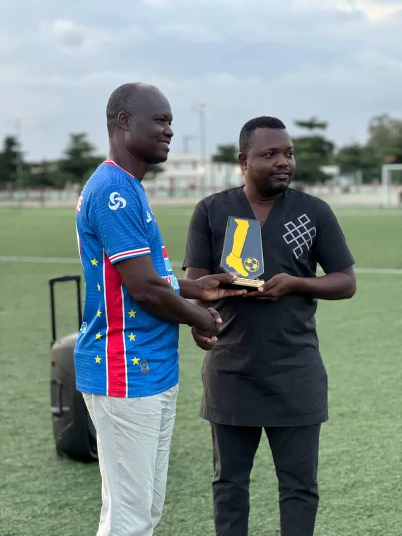 Deux hommes échangent une poignée de main, l’un tenant un trophée sur un terrain de football en plein air
