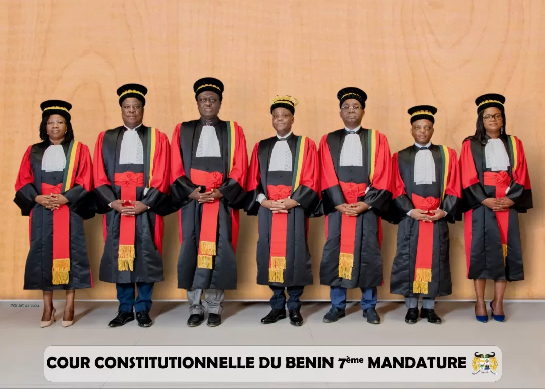 Sept membres vêtus de robes rouges et noires posant devant un fond en bois clair avec un panneau inscrit « COUR CONSTITUTIONNELLE DU BENIN 7ème MANDATURE