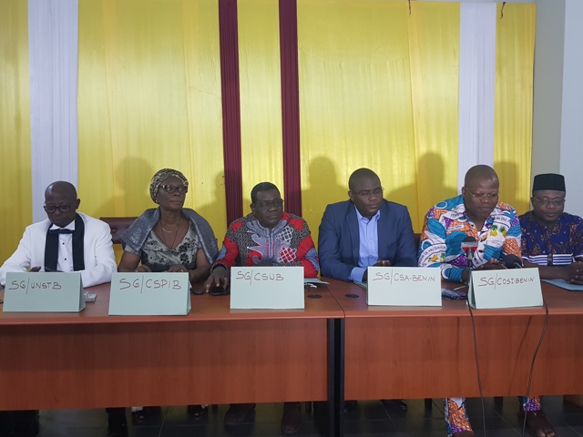 Six personnes assises derrière une table avec des pancartes indiquant des sigles devant elles et un rideau jaune en arrière-plan