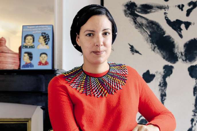 Femme portant un pull rouge et un collier coloré assise devant un mur décoré de peintures et d'objets d'art