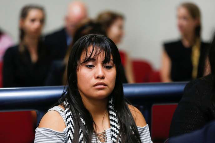 Jeune femme assise au premier plan dans une salle, regard pensif, plusieurs personnes floues en arrière-plan