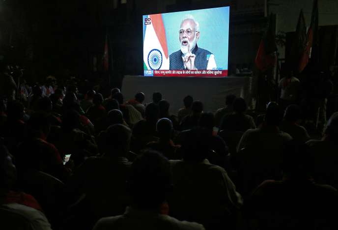 Des silhouettes de personnes regardant un homme parlant sur un grand écran avec un drapeau tricolore en arrière-plan