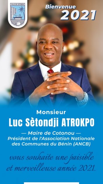 Homme en costume cravate souriant devant un fond flou avec texte de vœux pour l'année 2021 et blason de la ville de Cotonou