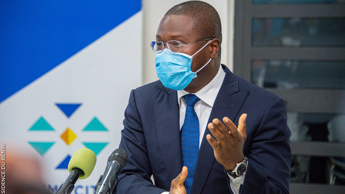Un homme en costume sombre, lunettes et masque bleu et blanc parle lors d une conférence avec deux micros devant lui