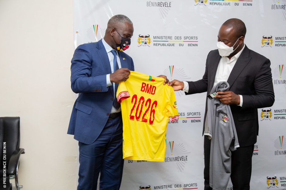 Deux hommes en costume et masque échangent des maillots devant un fond blanc marqué « Ministère des Sports République du Bénin