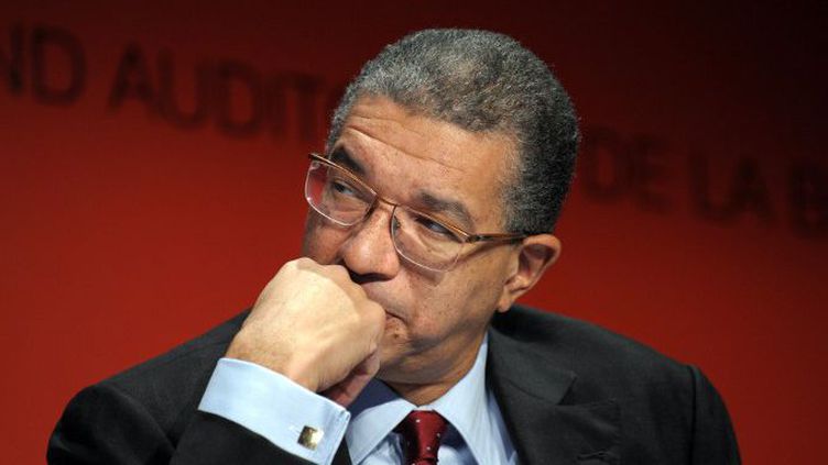 Homme mûr en costume sombre et chemise bleue, lunettes, pose sa main devant la bouche sur fond rouge flou