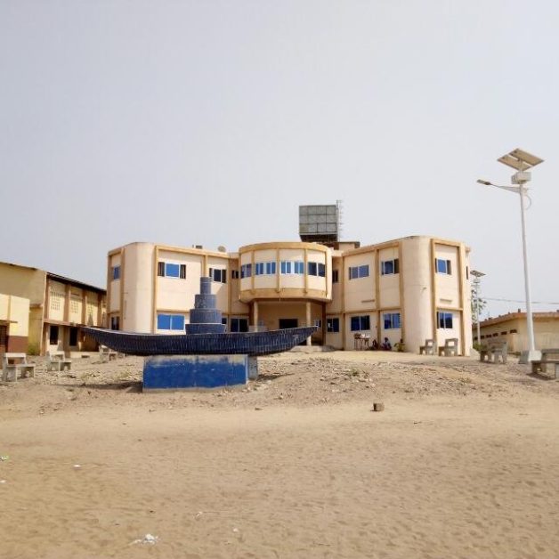 Bâtiment administratif beige sur terrain sablonneux avec une sculpture bleue au premier plan et un lampadaire solaire à droite