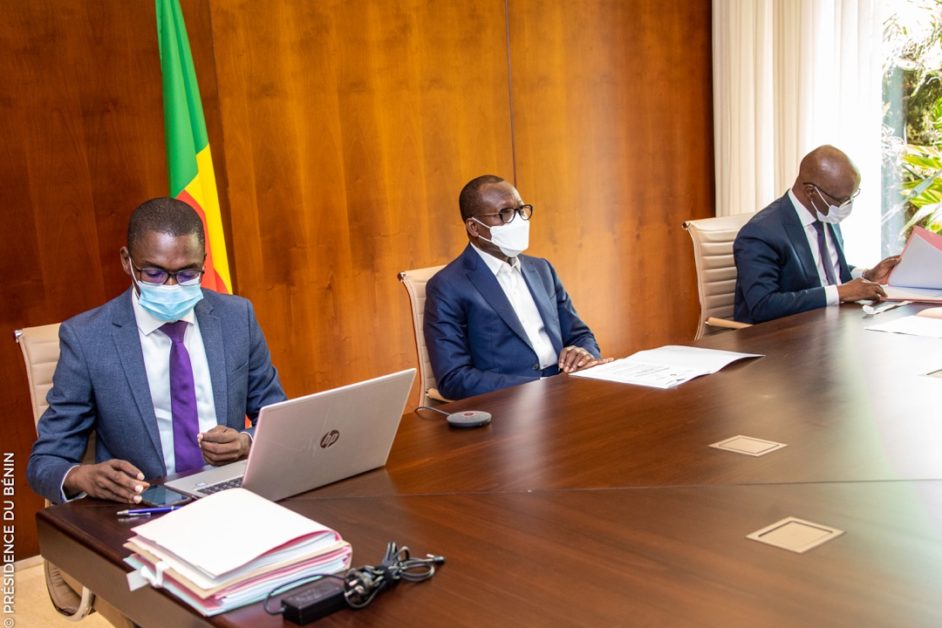 Trois hommes portant des masques assis à une grande table en bois avec des documents et un ordinateur portable devant eux