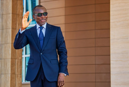 Homme en costume sombre, lunettes, levant la main droite en signe de salutation devant un mur beige et une porte bleue