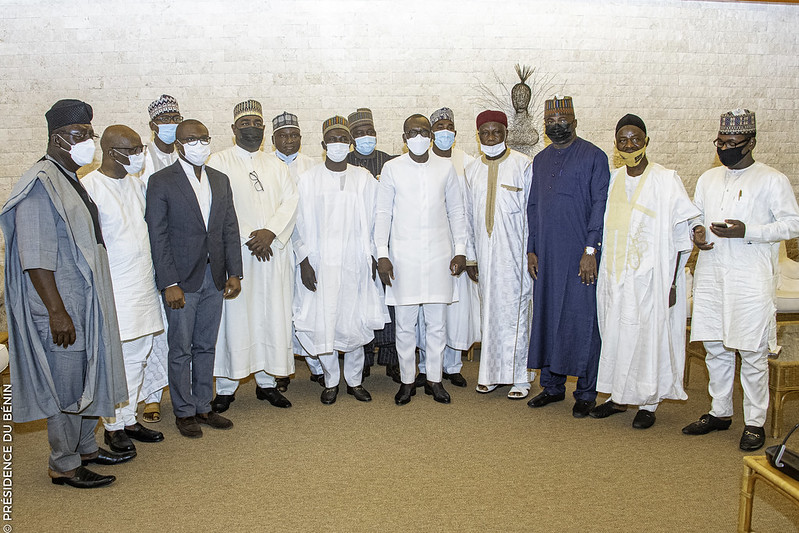 Groupe d’hommes en tenue traditionnelle africaine et costume occidental posant ensemble dans une pièce avec un mur en pierre claire