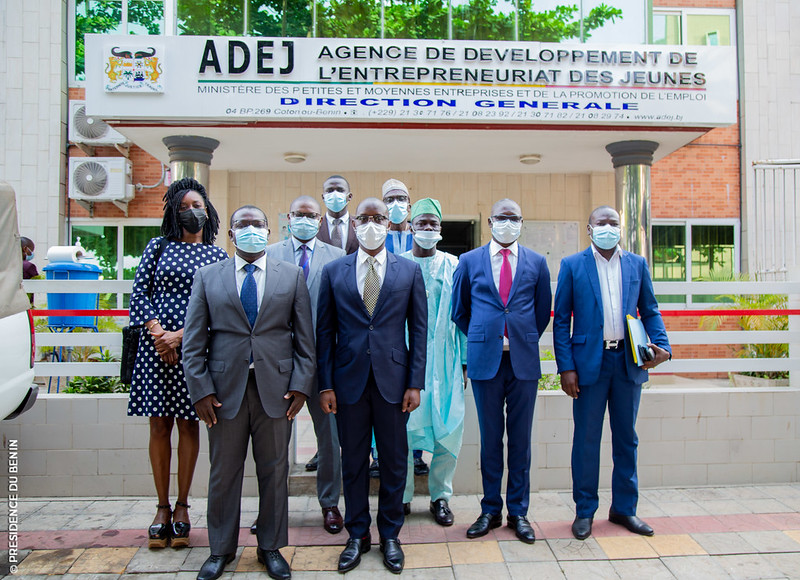Groupe de neuf personnes habillées formellement et portant des masques posant devant un bâtiment avec une enseigne officielle