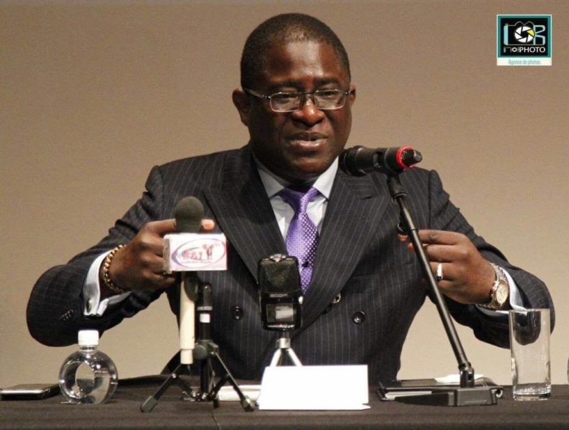 Homme en costume rayé et cravate prise la parole devant plusieurs micros lors d'une conférence assis à une table noire