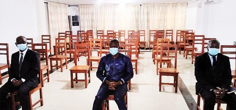 Trois hommes masqués assis espacés sur des chaises en bois dans une grande salle claire avec des rideaux beige fermeture.