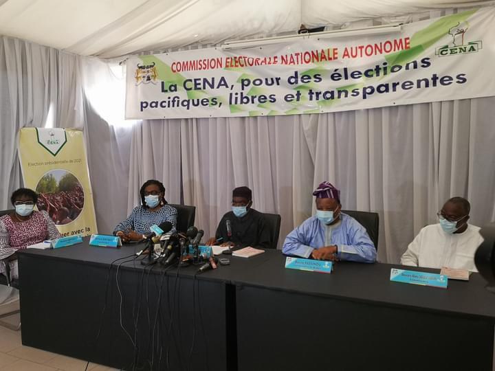 Cinq personnes assises derrière une table avec des micros sous une banderole sur des élections pacifiques libres et transparentes