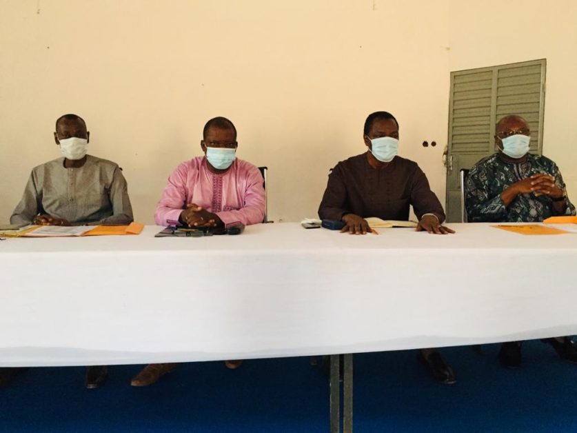 Quatre hommes assis à une table vêtus de chemises et portant des masques chirurgicaux dans une salle simple