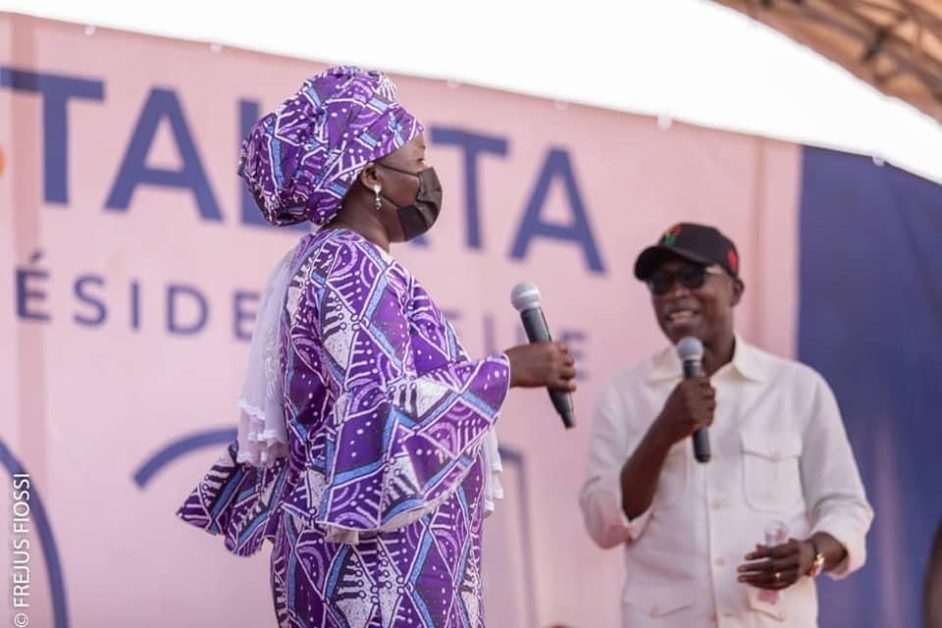 Une femme en vêtement traditionnel violet et un homme en chemise blanche tiennent chacun un micro sur une scène devant une bannière partiellement visible