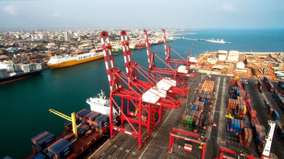Grand port maritime avec plusieurs grues rouges et conteneurs empilés le long du quai face à la mer et la ville en arrière-plan