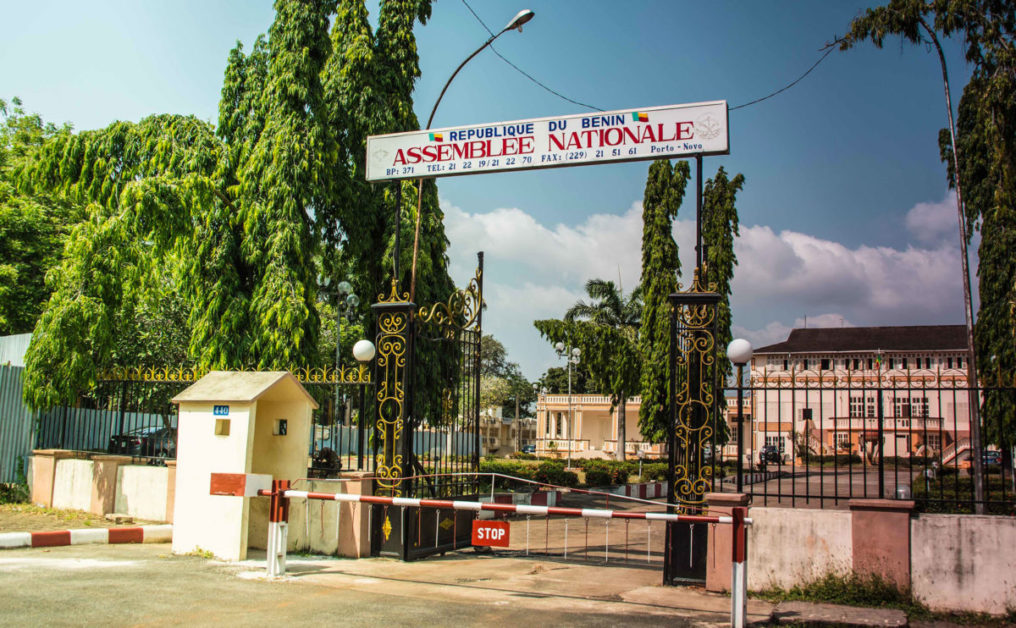Entrée principale d un bâtiment officiel entouré de plantations avec un portail métallique fermé et une barrière rouge et blanche