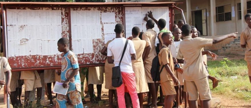 Des élèves consultent un grand tableau d'affichage en plein air devant un bâtiment scolaire en milieu urbain