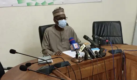 Homme masqué en tenue traditionnelle assis derrière une table avec plusieurs microphones lors d une conférence