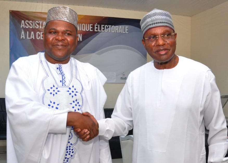 Deux hommes en tenue traditionnelle blanche se serrent la main devant une bannière partiellement visible sur fond bleu