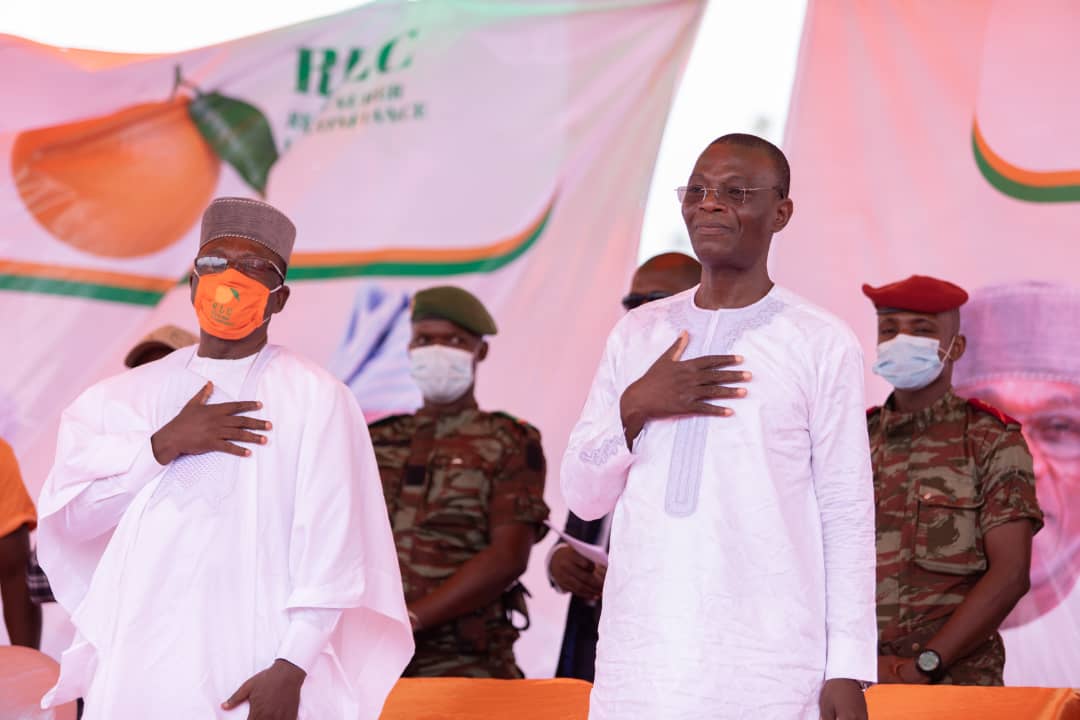 Deux hommes habillés en tenue traditionnelle debout main sur le cœur devant des militaires en uniformes avec masques faciaux