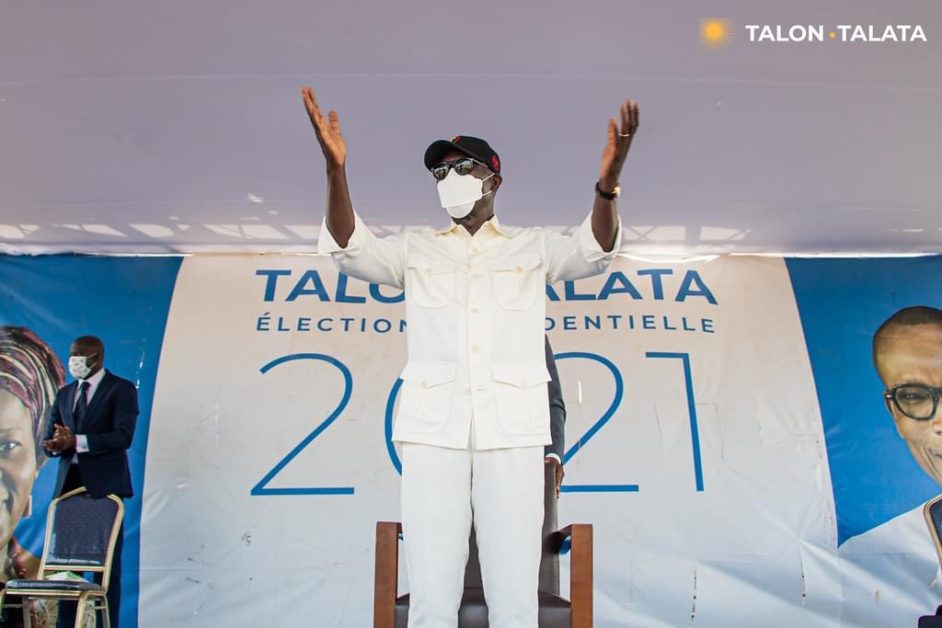 Un homme en tenue blanche, casquette et masque, lève les bras devant une banderole électorale 2021 pendant un discours