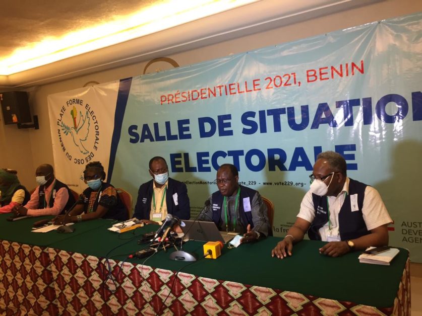 Cinq personnes assises à une table verte devant une grande bâche indiquant salle de situation électorale