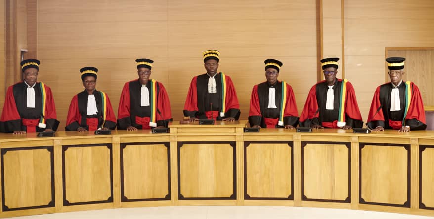 Sept membres en tenue officielle rouge et noire debout derrière un bureau en bois clair dans une salle officielle