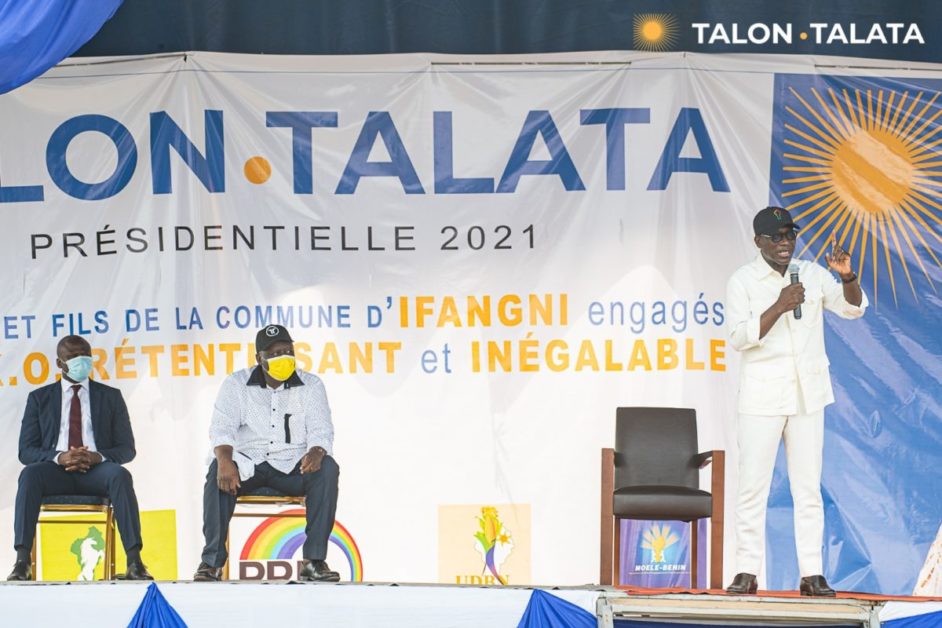 Trois hommes sur scène dont un debout parlant devant une grande banderole électorale aux couleurs bleu, blanc et orange