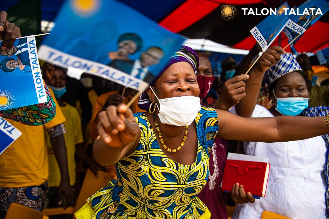 Des personnes masquées agitent des drapeaux aux couleurs bleue, jaune et blanche lors d’un rassemblement politique en plein air
