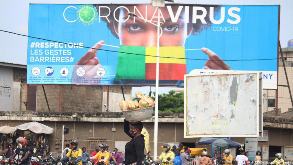 Foule dense devant un grand panneau sur le coronavirus avec un visage masqué aux couleurs d’un drapeau et une femme portant un panier sur la tête