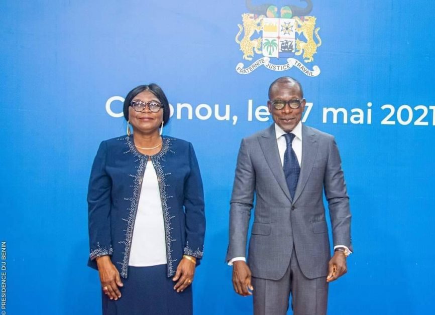 Une femme en tailleur bleu et un homme en costume gris posent devant un mur bleu portant un blason et un texte dates